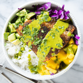 blackened salmon in a white bowl with rice, purple cabbage, mango, avocado and a green sauce sitting on a marble surface