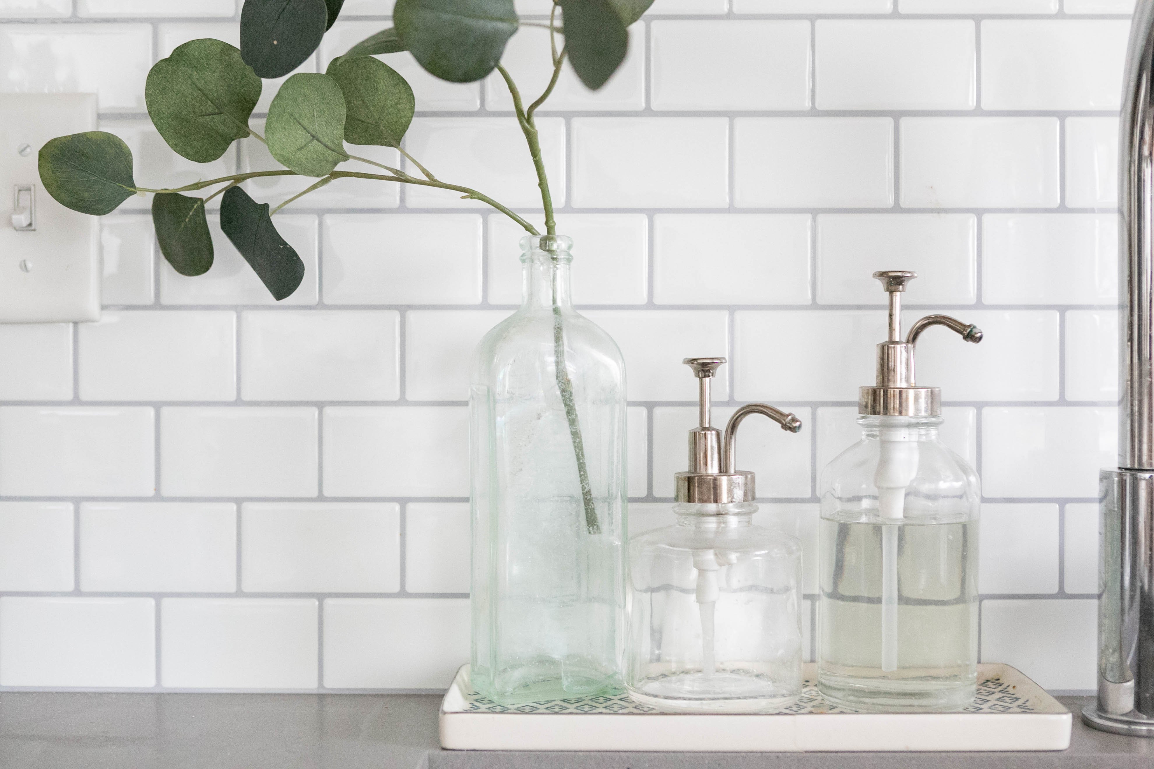 glass bottles with clear soap in a kitchen next to a clear jar with a green plant in it