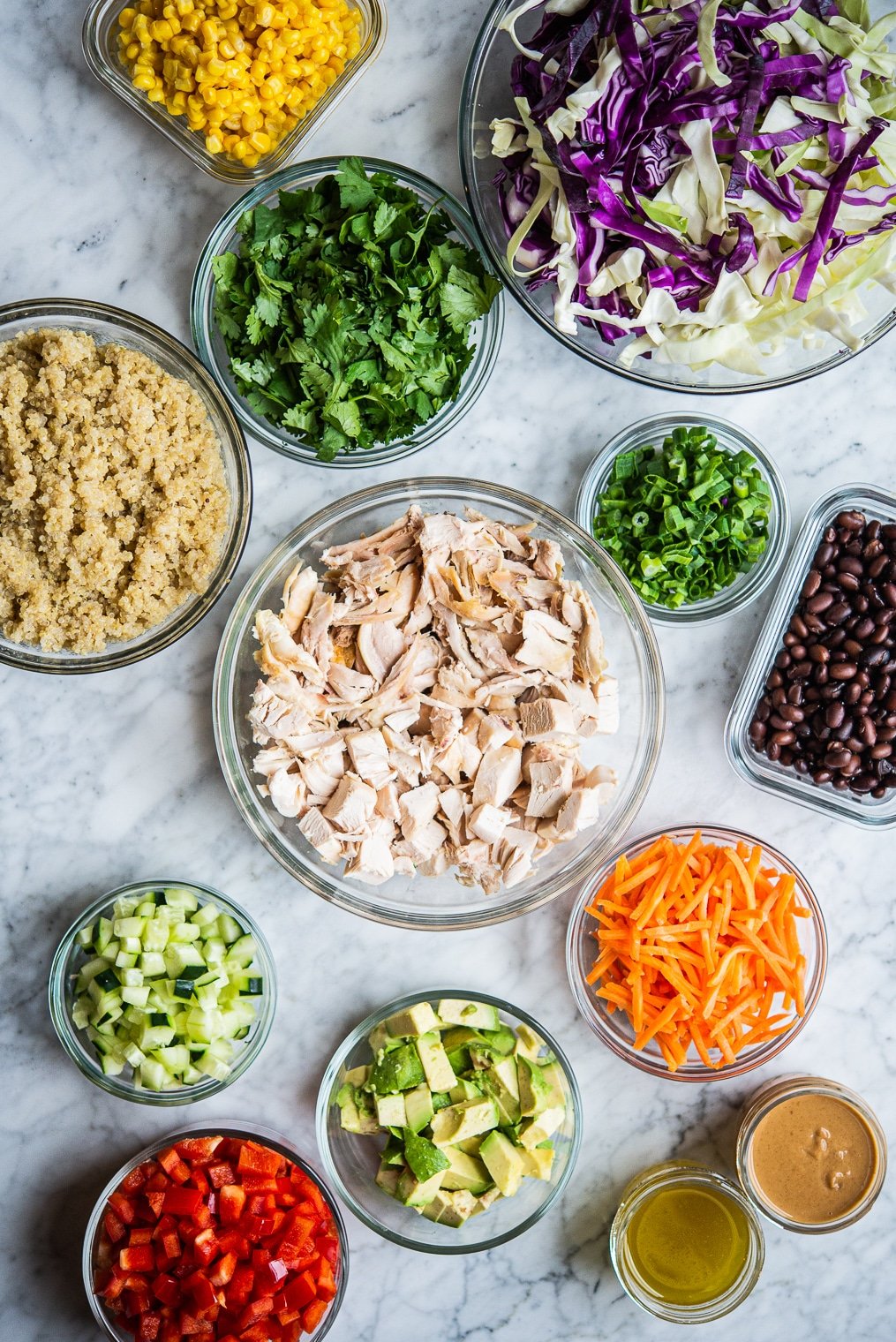 meal prep ingredients for thai chicken salad and southwestern quinoa salad in separate clear containers on a marble surface