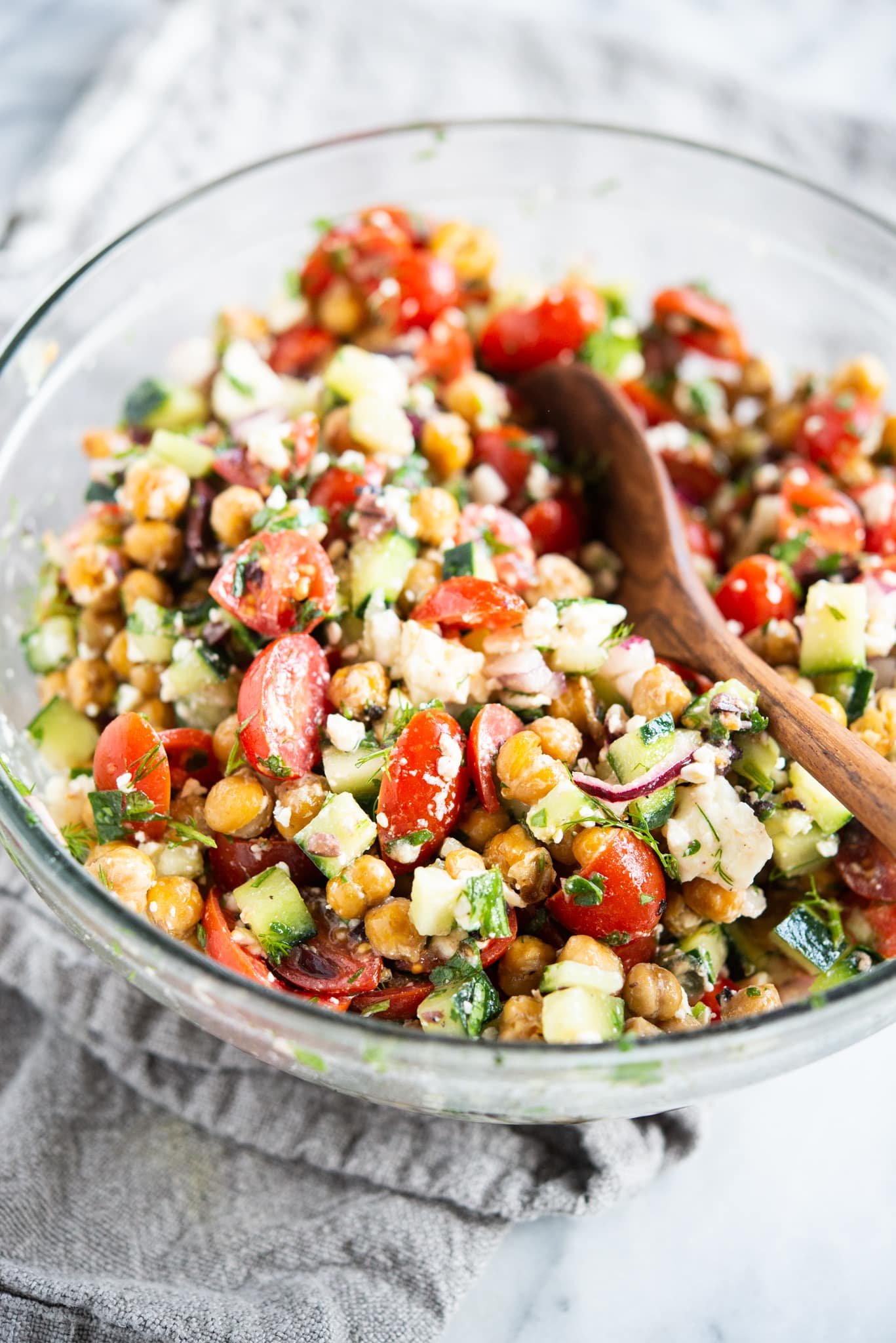 mediterranean chickpea salad - chickpeas, feta cheese, tomatoes, and cucumber - in a glass bowl on a marble surface with a wooden spoon inside