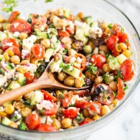 mediterranean chickpea salad - chickpeas, feta cheese, tomatoes, and cucumber - in a glass bowl on a marble surface with a wooden spoon inside