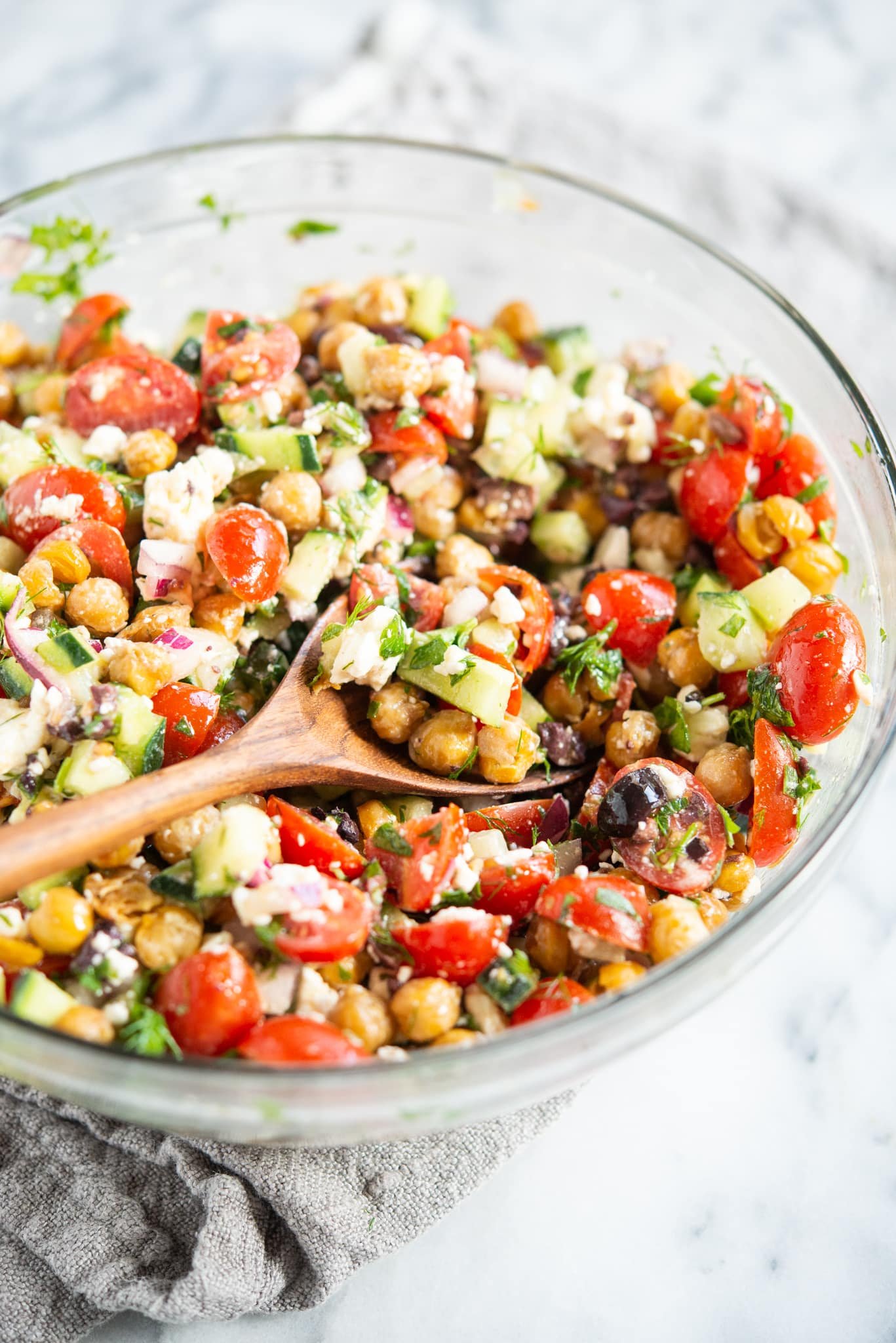 mediterranean chickpea salad - chickpeas, feta cheese, tomatoes, and cucumber - in a glass bowl on a marble surface with a wooden spoon inside