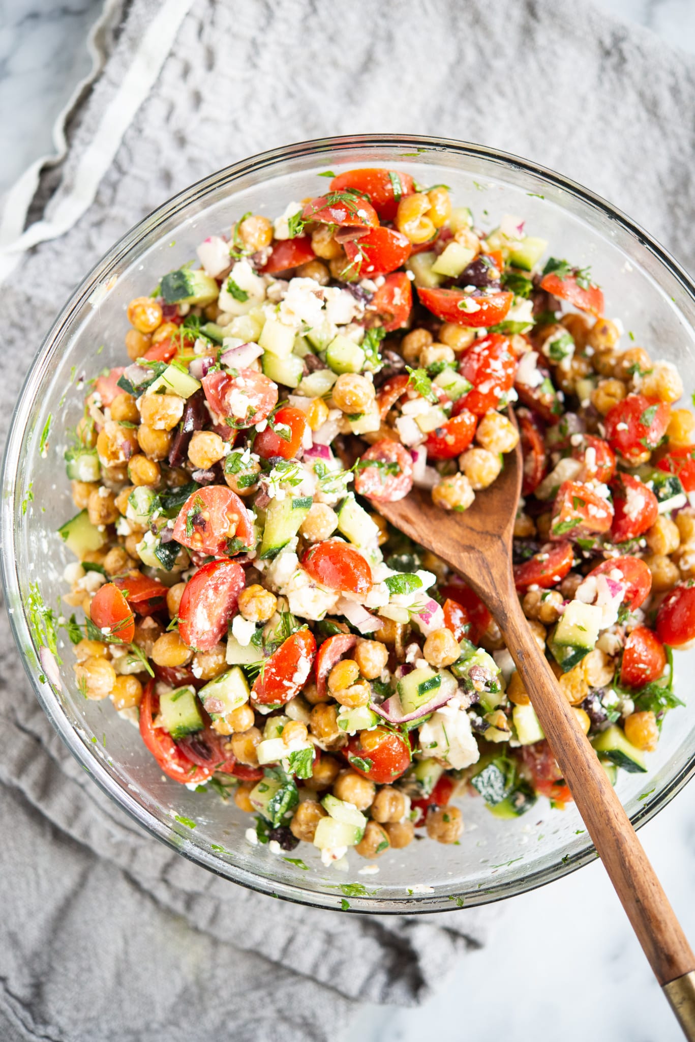 mediterranean chickpea salad - chickpeas, feta cheese, tomatoes, and cucumber - in a glass bowl on a marble surface with a wooden spoon inside