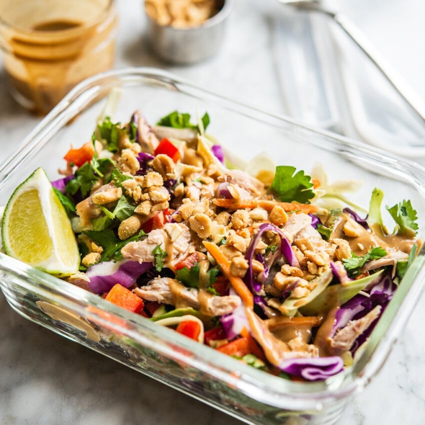 colorful thai chopped chicken salad in an individual serving glass meal prep container with peanut dressing + crushed peanuts in the background