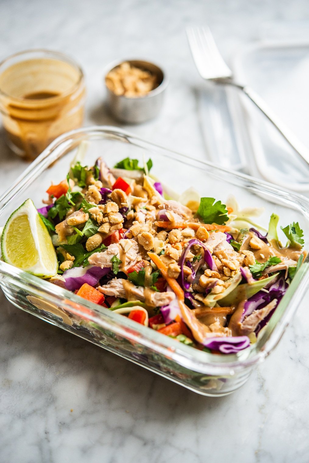 colorful thai chopped chicken salad in an individual serving glass meal prep container with peanut dressing + crushed peanuts in the background