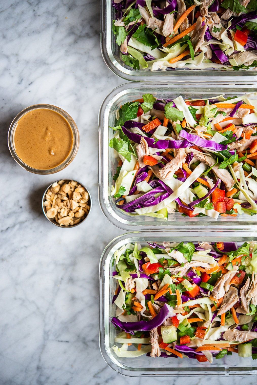 colorful thai chopped chicken salad in 3 individual serving glass meal prep containers sitting next to peanut dressing + crushed peanuts