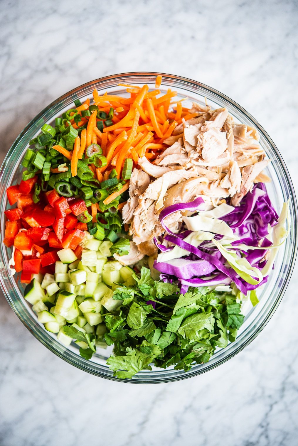 thai chopped chicken salad ingredients: carrots, cucumber, bell pepper, cabbage, chicken, and cilantro in a large glass bowl on a marble surface