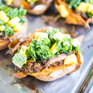 sweet potatoes stuffed with shredded jerk pork topped with kale and mangoes on a stainless steel sheet pan