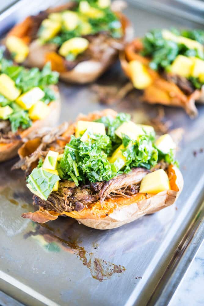 sweet potatoes stuffed with shredded jerk pork topped with kale and mangoes on a stainless steel sheet pan