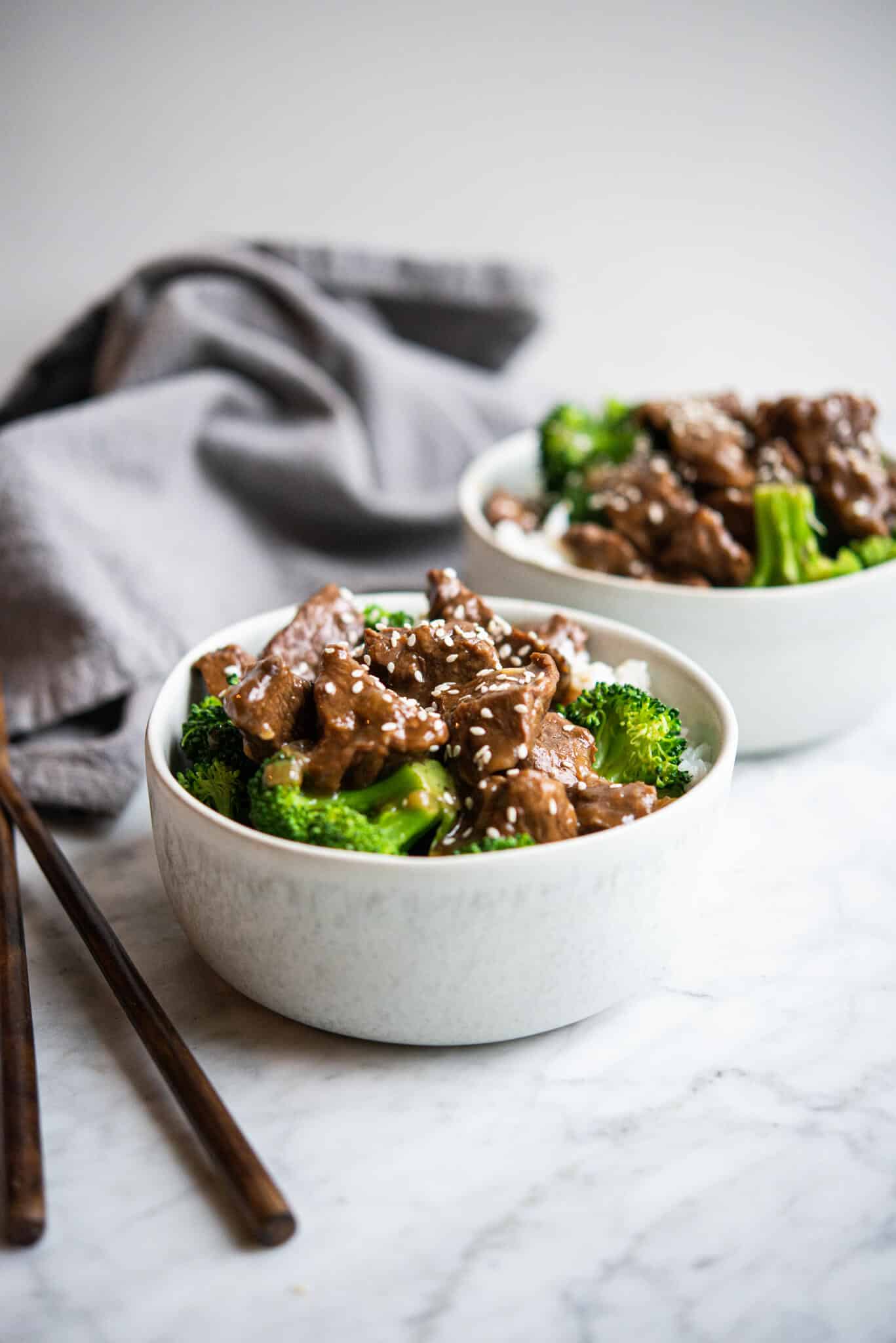 beef and broccoli over white rice in a white bowl on a marble surface with another bowl behind it
