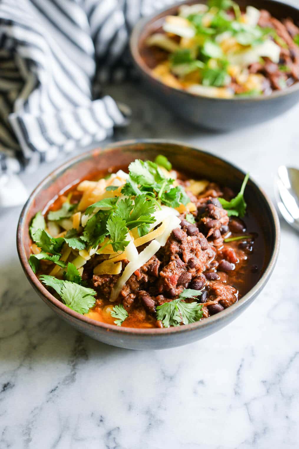 two bowls of slow cooker chili topped with shredded cheddar cheese and cilantro sitting on a marble surface