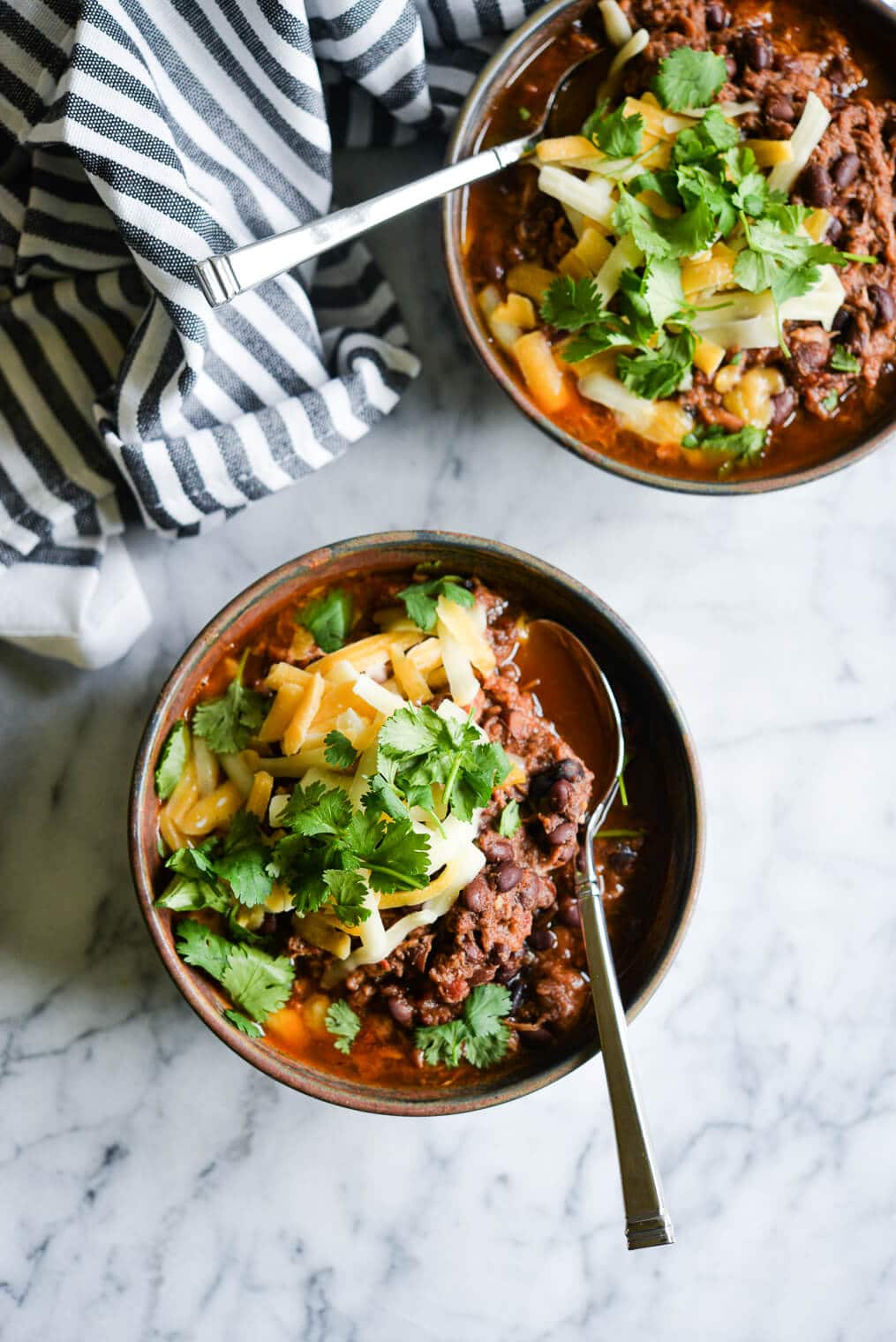 two bowls of beef and bean chili topped with shredded cheddar cheese and cilantro sitting on a marble surface