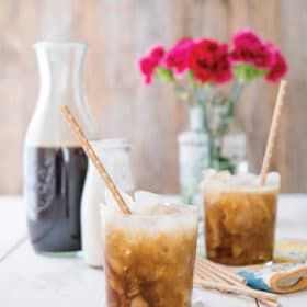 two cups of cold brew coffee with with paper straws in front of a wooden background with a pitcher of cold brew, cream, and pink flowers