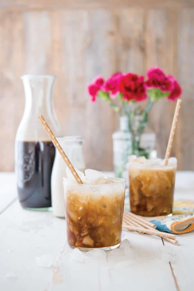 two cups of cold brew coffee with with paper straws in front of a wooden background with a pitcher of cold brew, cream, and pink flowers