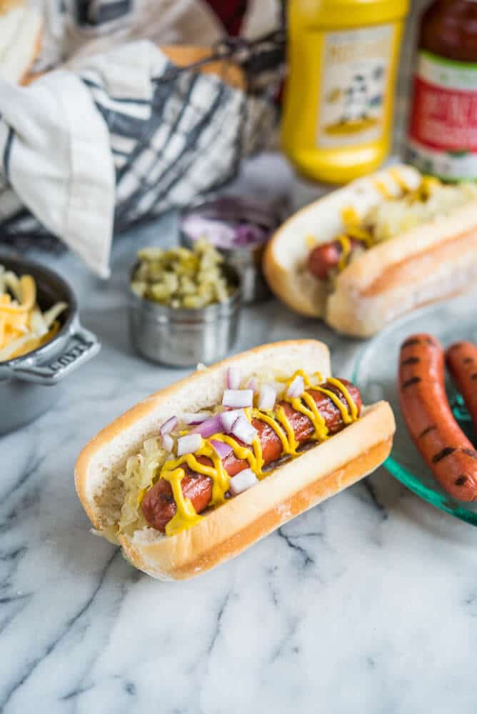 two hot dogs with sauerkraut, red onions, and mustard next to other toppings on a marble surface