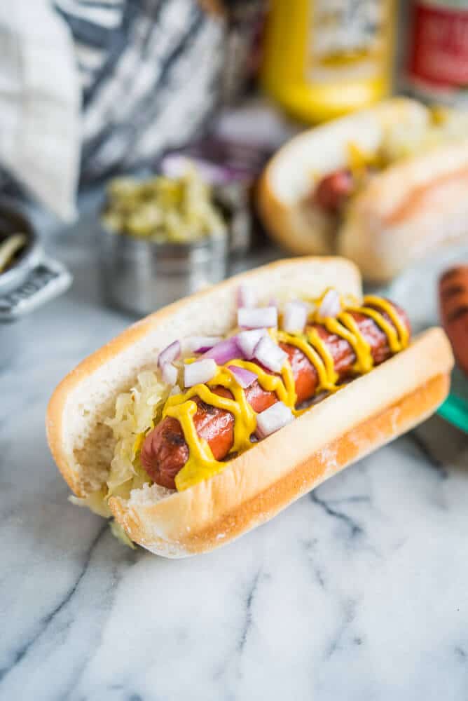 two hot dogs with sauerkraut, red onions, and mustard next to other toppings on a marble surface