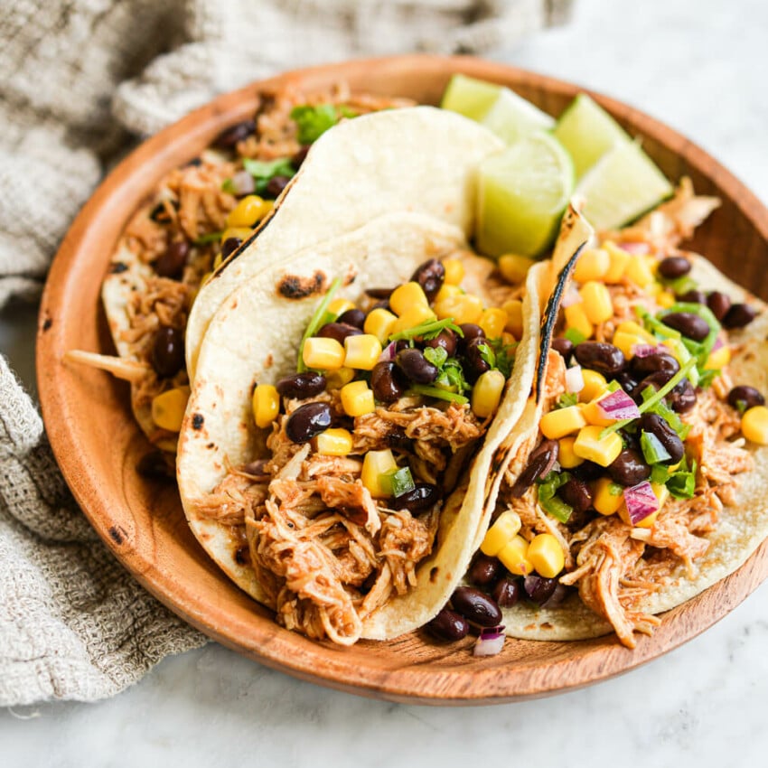 three instant pot bbq chicken tacos topped with black bean and corn salsa on a wooden plate on a marble surface
