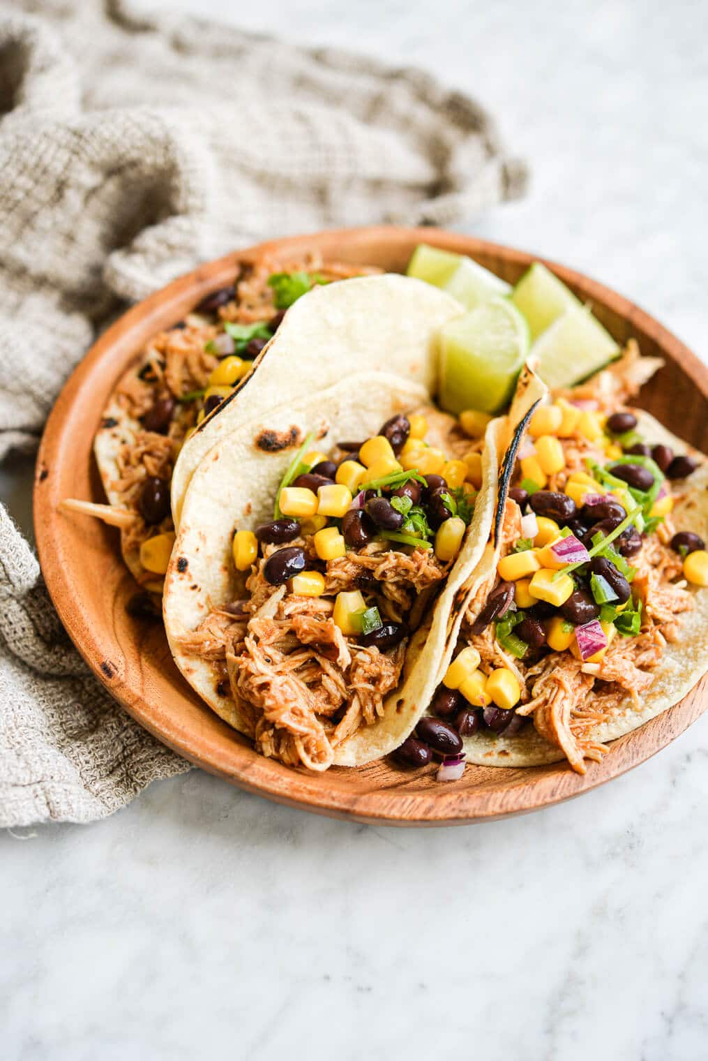 three instant pot bbq chicken tacos topped with black bean and corn salsa on a wooden plate on a marble surface