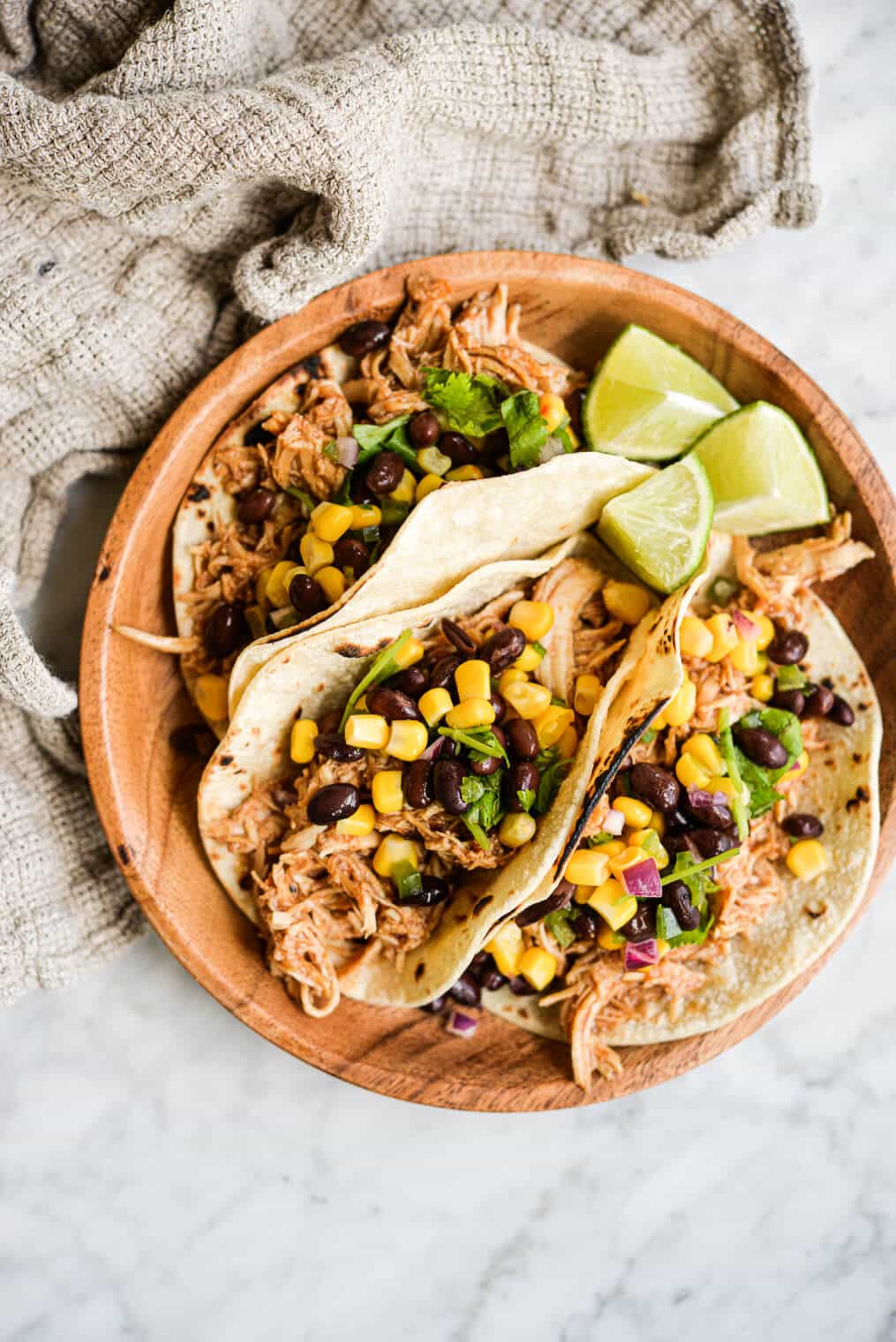 three instant pot bbq chicken tacos topped with black bean and corn salsa on a wooden plate on a marble surface