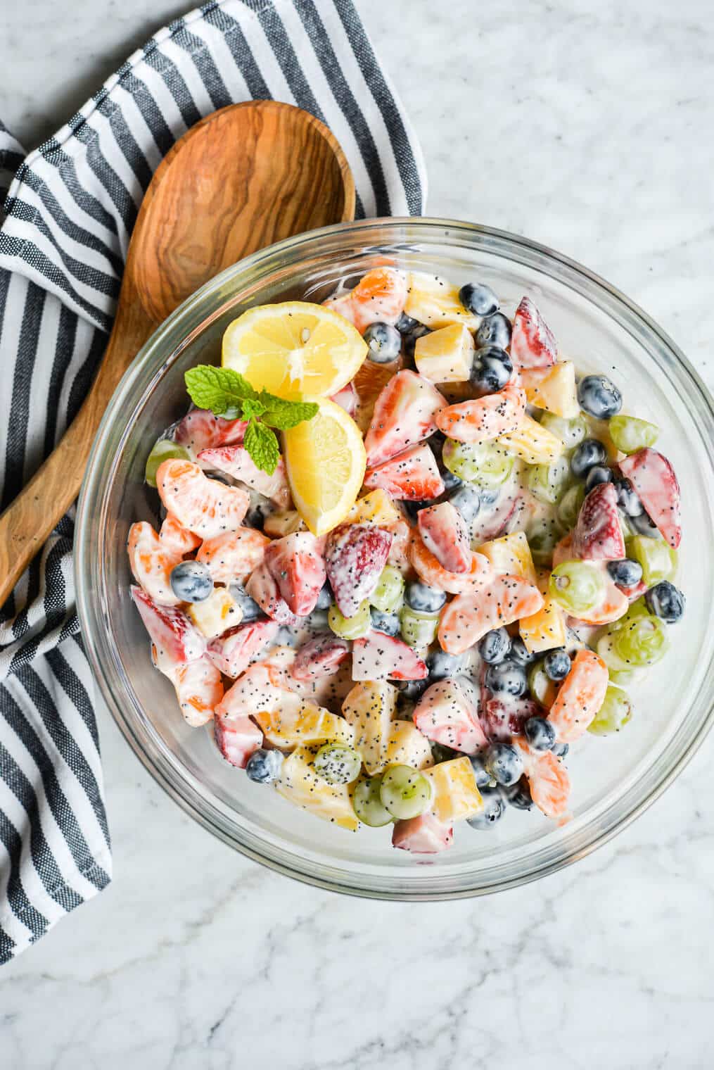 ready to eat yogurt fruit salad in a large glass bowl next to a wooden spoon and striped kitchen towel on a marble surface