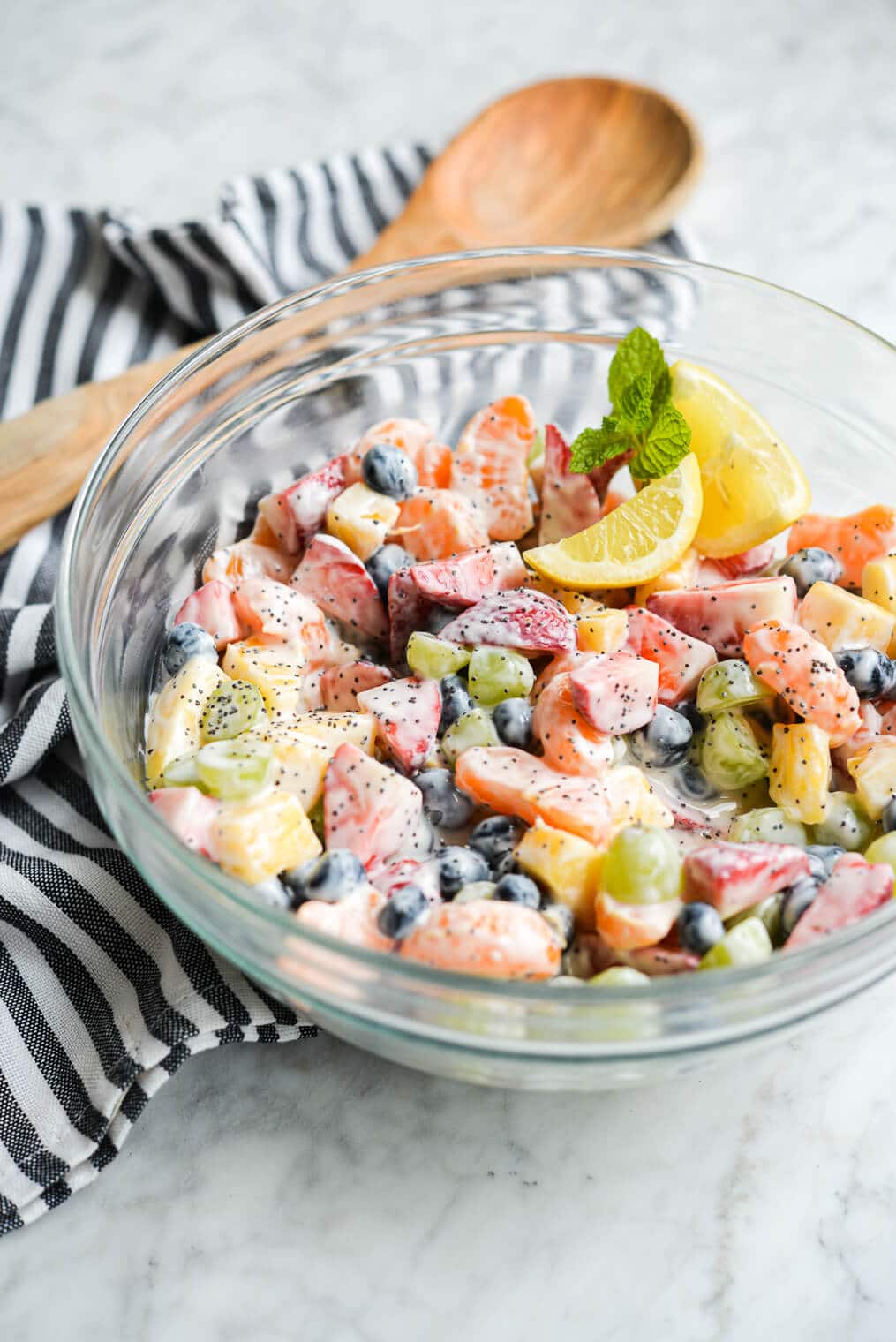 ready to eat yogurt fruit salad in a large glass bowl next to a wooden spoon and striped kitchen towel on a marble surface