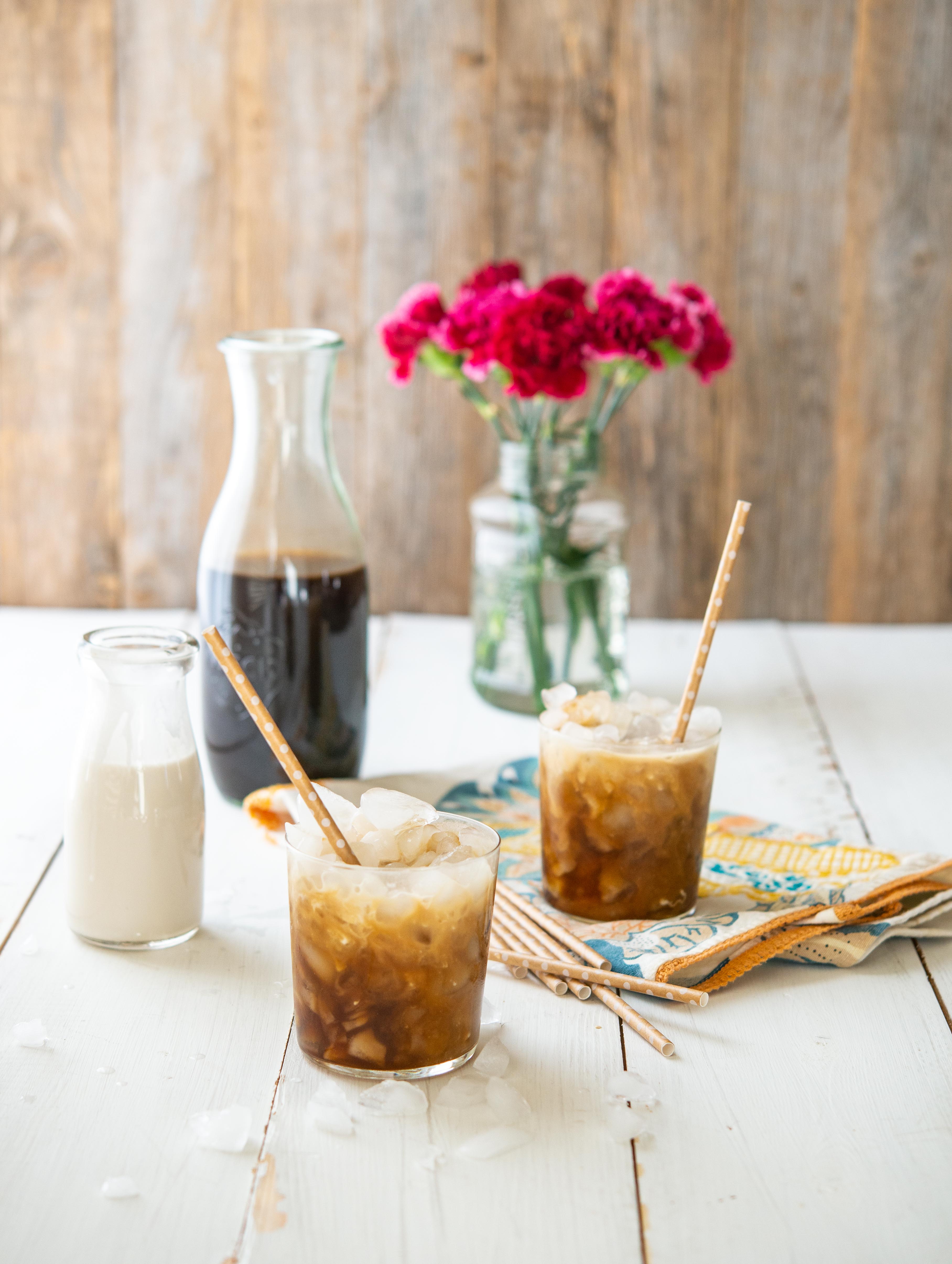 two cups of cold brew coffee with with paper straws in front of a wooden background with a pitcher of cold brew, cream, and pink flowers