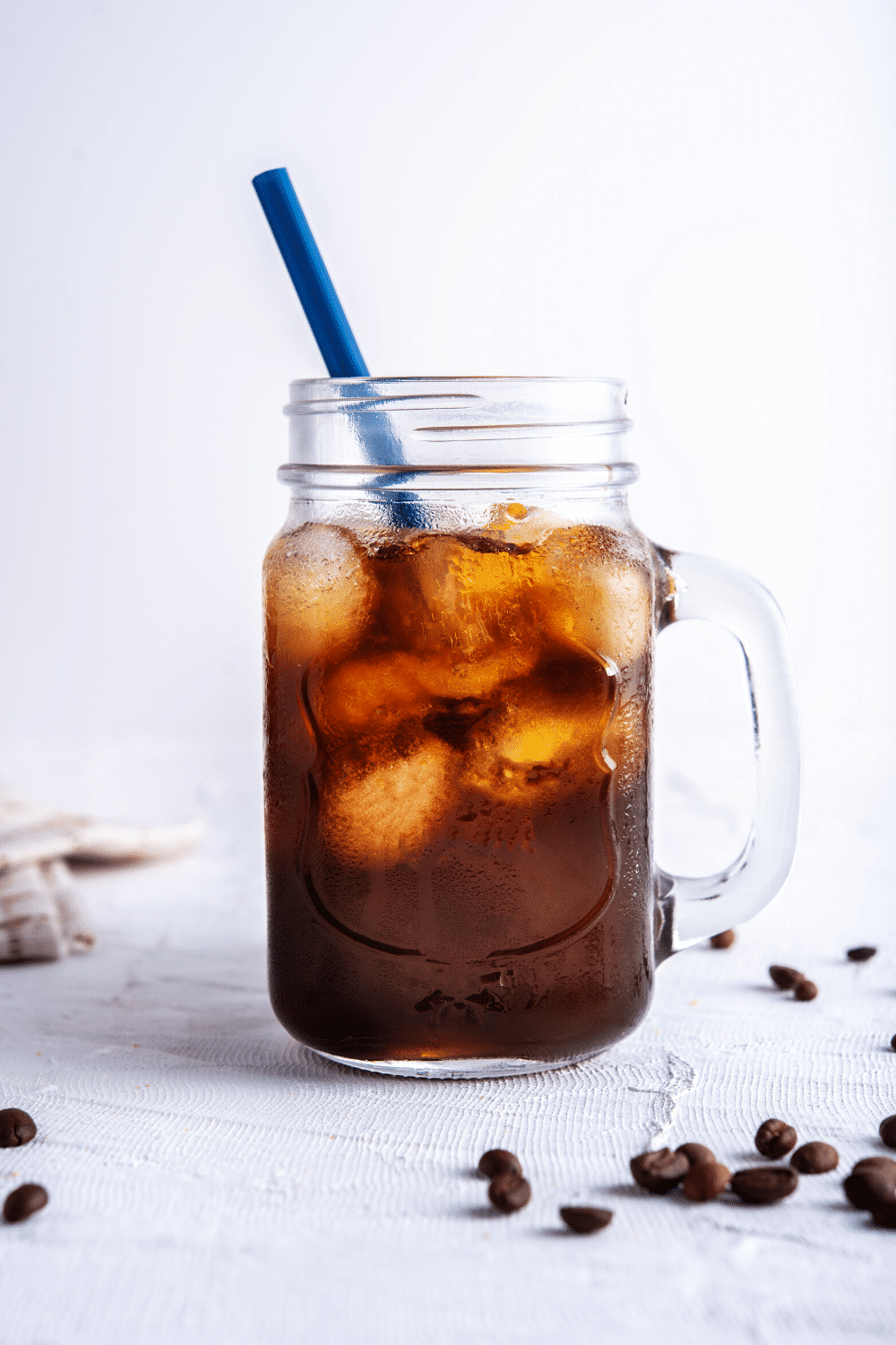 Mason jar mug with cold brewed coffee and a blue straw with coffee beans scattered on the table