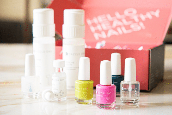 dazzle dry nail kit with three nail polishes, a top coat, and a remover, in front of a bright pink box