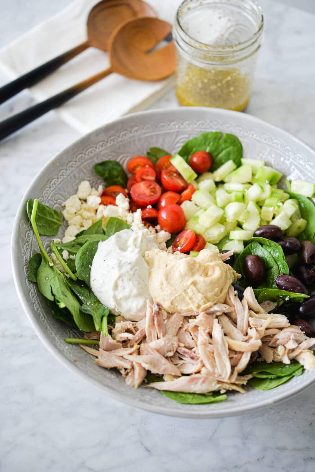 a bed of spinach topped with halved tomatoes, feta cheese, cucumbers, olives, shredded chicken, hummus, and tzatziki sitting next to a jar of homemade greek dressing and two wooden serving spoons all on a marble surface