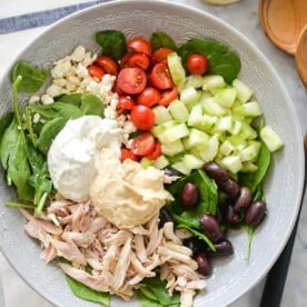 a bed of spinach topped with halved tomatoes, feta cheese, cucumbers, olives, shredded chicken, hummus, and tzatziki sitting next to a jar of homemade greek dressing and two wooden serving spoons all on a marble surface