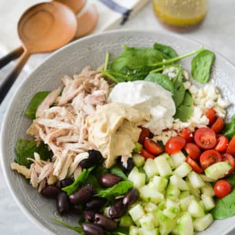 a bed of spinach topped with halved tomatoes, feta cheese, cucumbers, olives, shredded chicken, hummus, and tzatziki sitting next to a jar of homemade greek dressing and two wooden serving spoons all on a marble surface