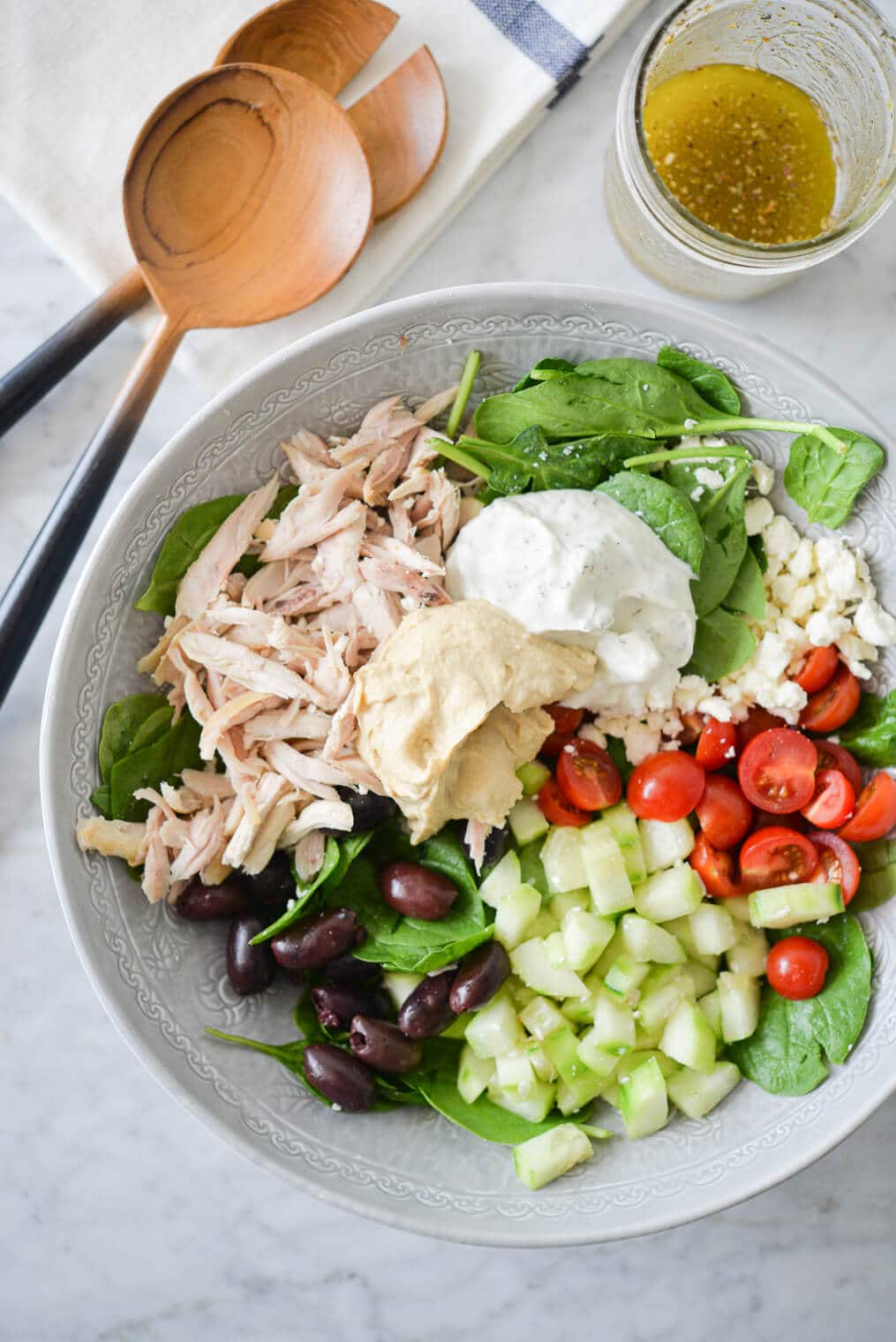 a bed of spinach topped with halved tomatoes, feta cheese, cucumbers, olives, shredded chicken, hummus, and tzatziki sitting next to a jar of homemade greek dressing and two wooden serving spoons all on a marble surface