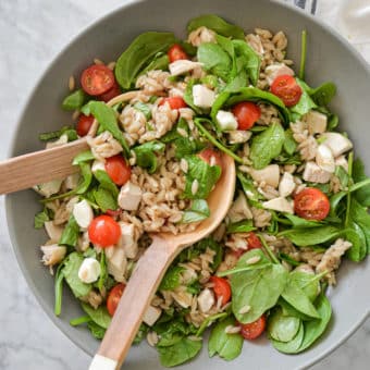 a large gray bowl filled with orzo, cherry tomatoes, spinach, mozzarella pearls, and chicken with two wooden serving spoons on a marble surface