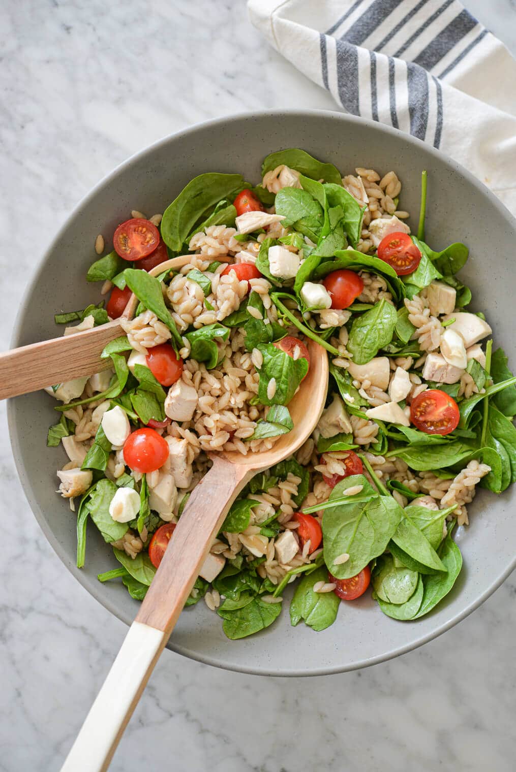 a large gray bowl filled with orzo, cherry tomatoes, spinach, mozzarella pearls, and chicken with two wooden serving spoons on a marble surface