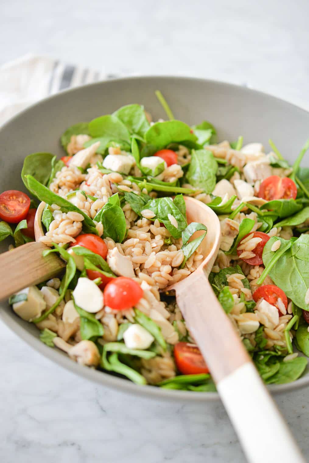 a large gray bowl filled with orzo, cherry tomatoes, spinach, mozzarella pearls, and chicken with two wooden serving spoons on a marble surface