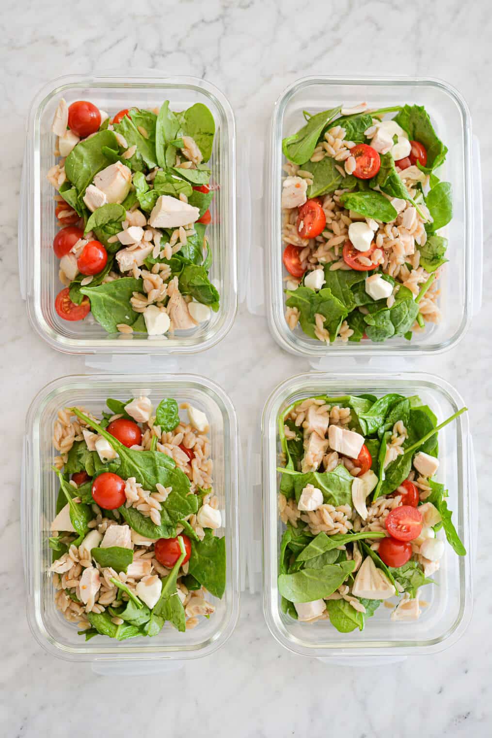 4 rectangular glass meal prep containers filled with orzo caprese salad sitting on a marble surface