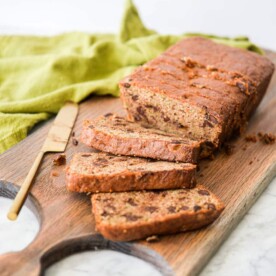 a batch of sliced chocolate chip zucchini bread laying on a wooden cutting board next to a brass butter knife on a marble surface