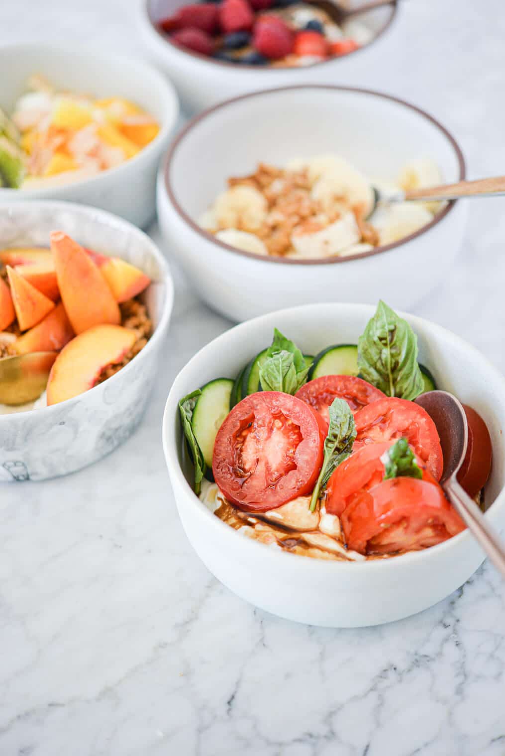 a white bowl filled with plain Greek yogurt, sliced cucumbers and tomatoes, fresh basil leaves, and balsamic vinegar surrounded by various other Greek yogurt bowls all sitting on a marble surface