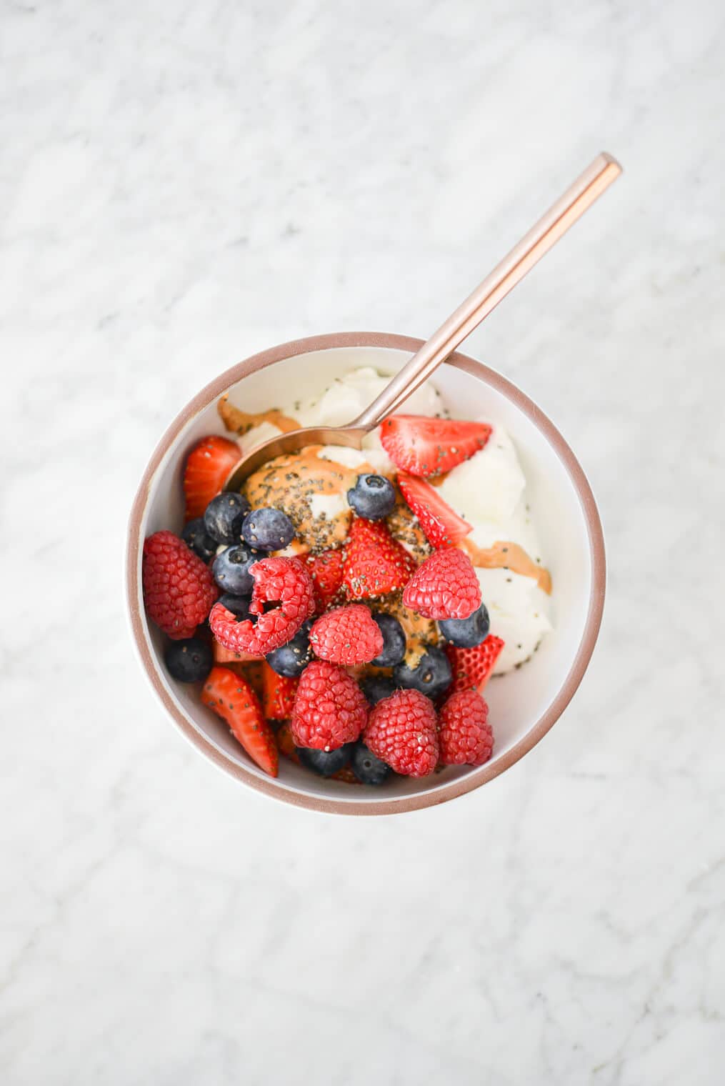 top view of a white bowl filled with plain Greek yogurt, raspberries, strawberries, blueberries, chia seeds, and cashew butter sitting on a marble surface