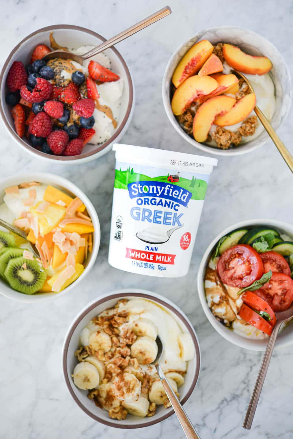 Top view of 5 Greek yogurt bowls (tropical, berry, peaches and cream, banana nut, and cucumber caprese) surrounding a quart of Stonyfield organic plain Greek yogurt on a marble surface