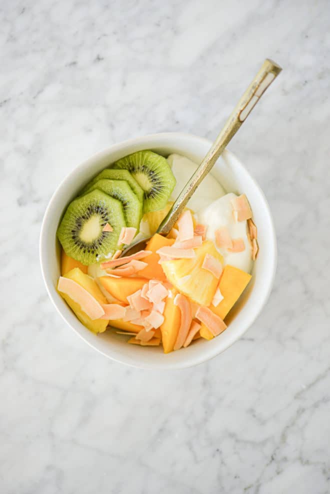 top view of a tropical Greek yogurt bowl loaded with sliced kiwi, mango, pineapple, and toasted coconut sitting on a marble surface