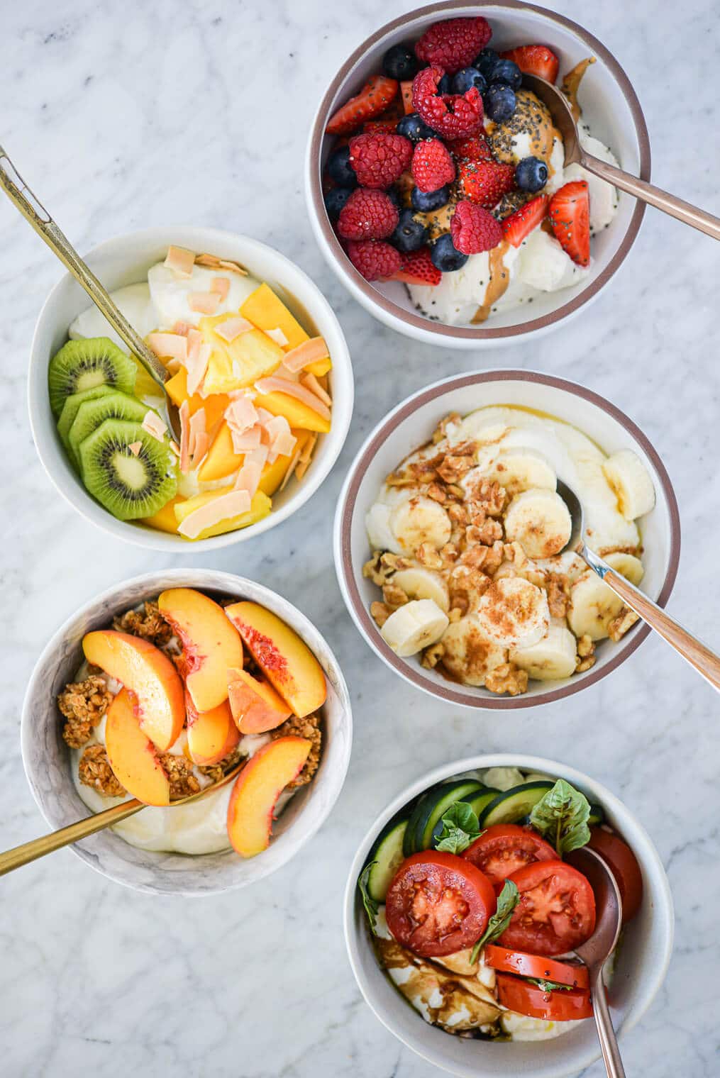 Top view of 5 Greek yogurt bowls (tropical, berry, peaches and cream, banana nut, and cucumber caprese) on a marble surface