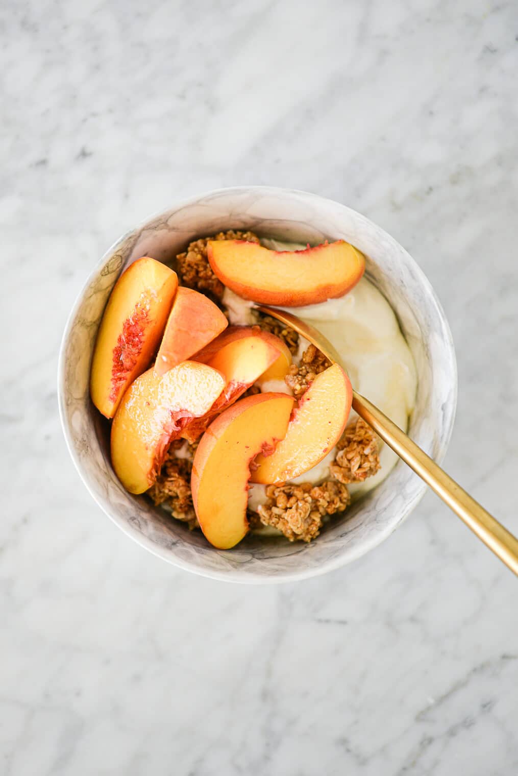 top view of a marble bowl with plain Greek yogurt, sliced fresh peaches, granola, and honey on a marble surface