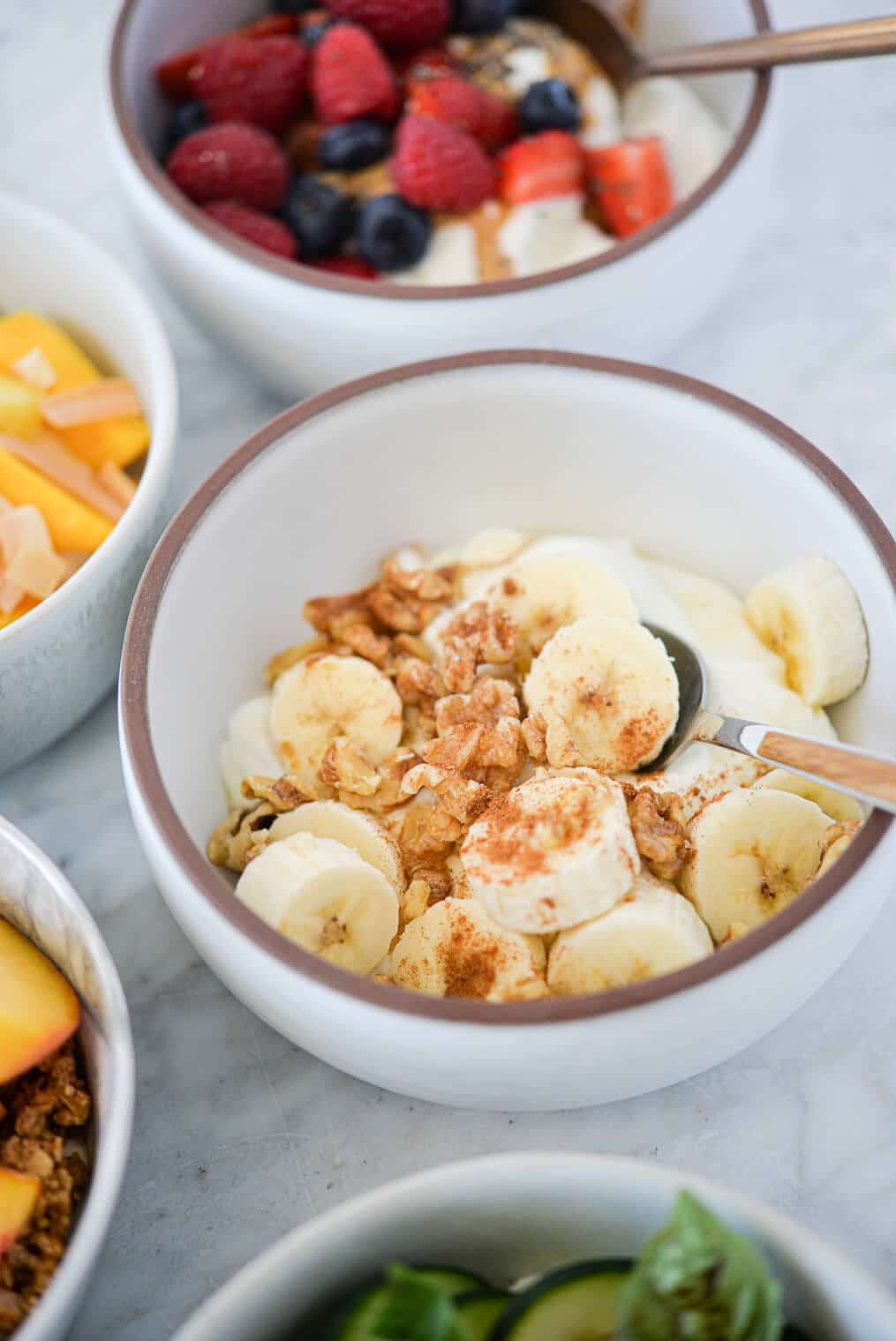 a white bowl with plain Greek yogurt, chopped bananas, walnuts, and cinnamon next to several other Greek yogurt bowls all on a marble surface