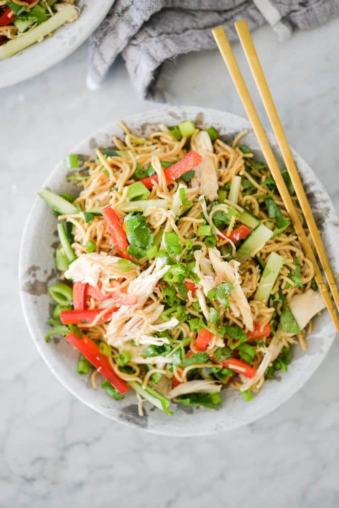 top view of sesame noodles with raw strips of cucumber, red bell peppers, and snap peas in a stone bowl with gold chopsticks resting on the rim all on a marble surface