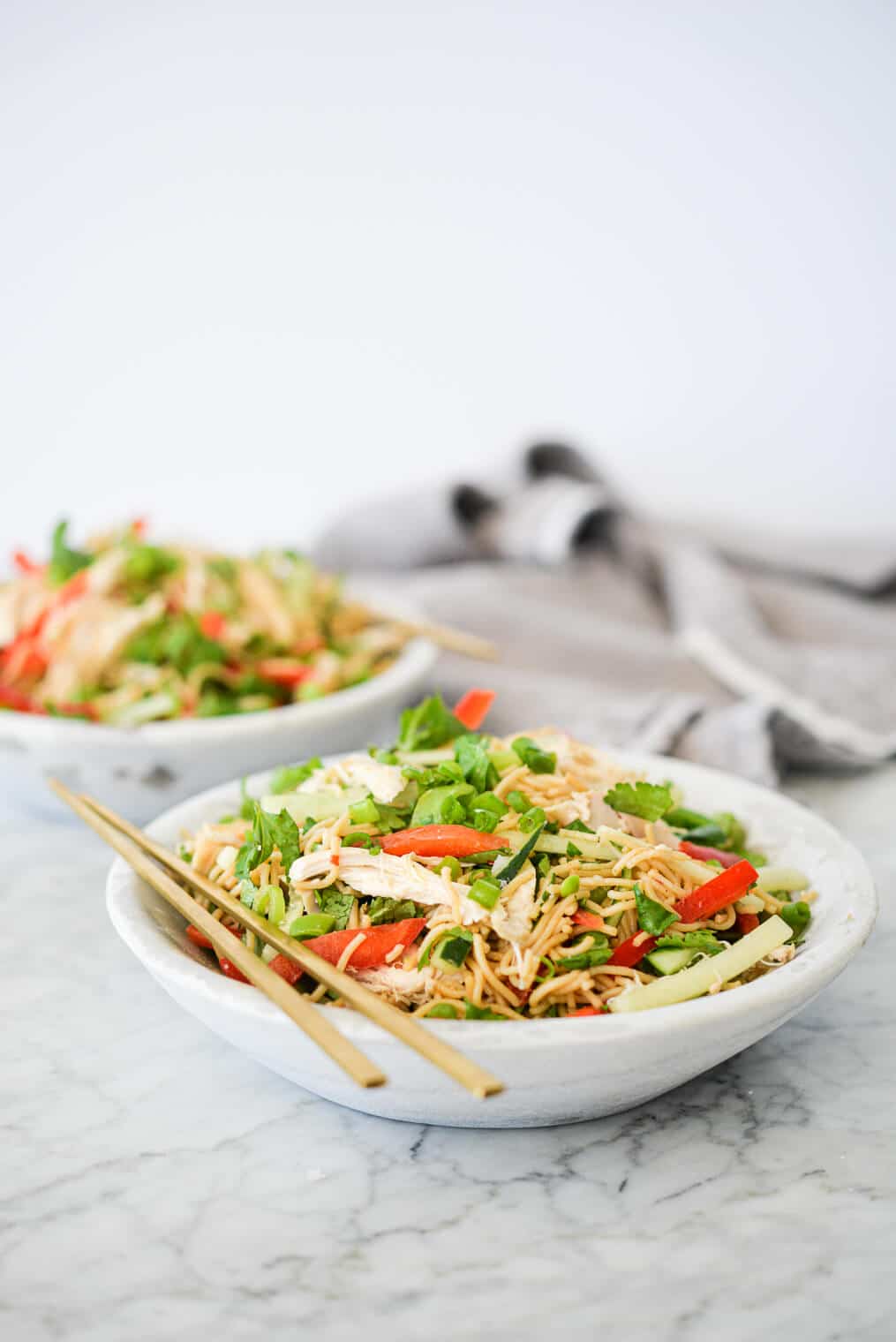 side view of two bowls of sesame noodles with raw strips of cucumber, red bell peppers, and snap peas in a stone bowl with gold chopsticks resting on the rim all on a marble surface