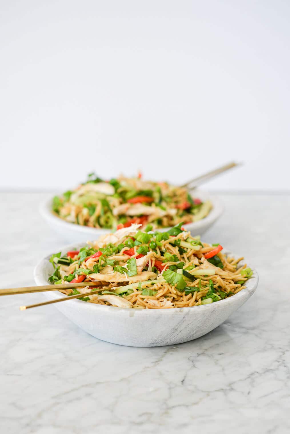 side view of two bowls of sesame noodles with raw strips of cucumber, red bell peppers, and snap peas in a stone bowl with gold chopsticks resting on the rim all on a marble surface