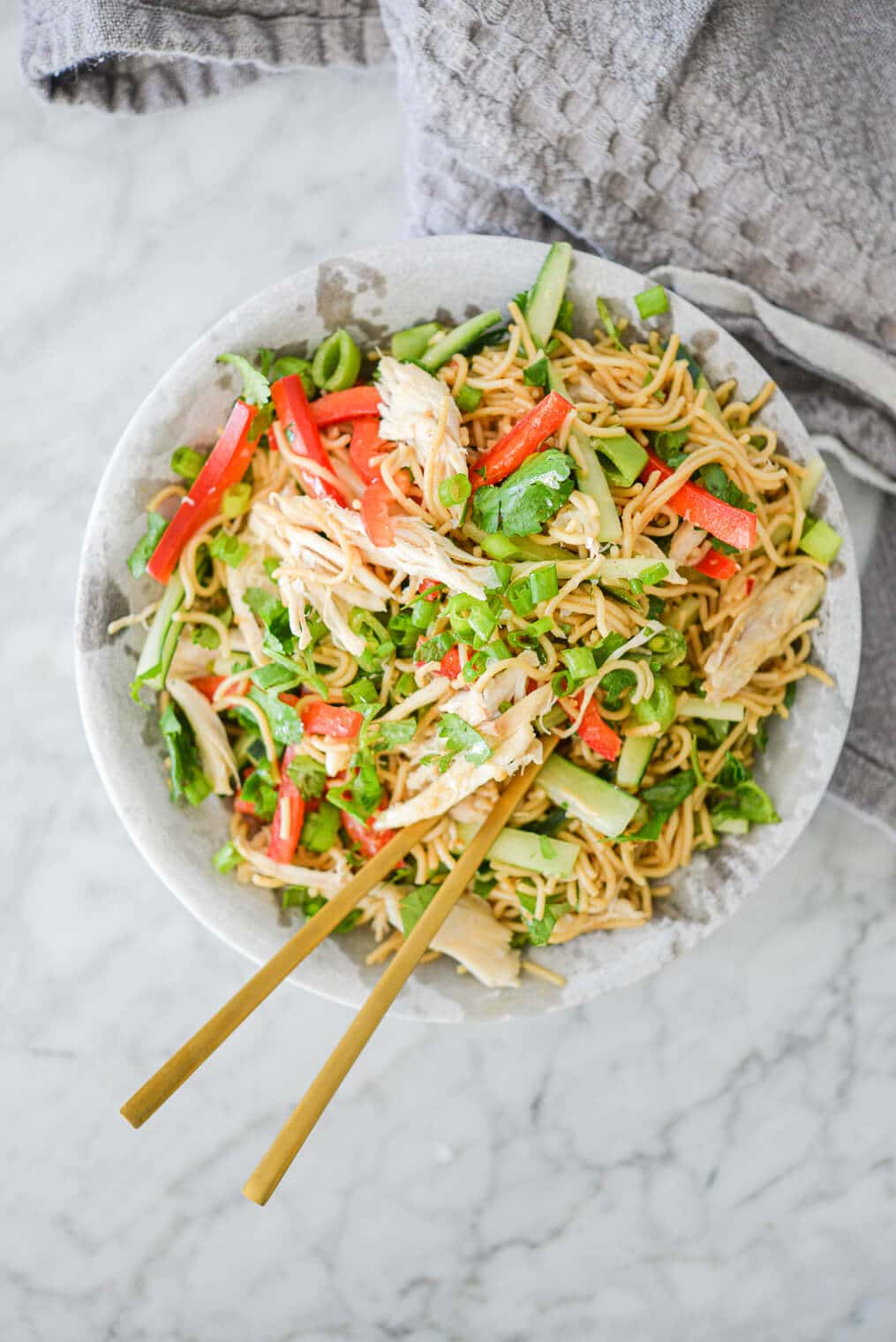 top view of sesame noodles with raw strips of cucumber, red bell peppers, and snap peas in a stone bowl with gold chopsticks resting on the rim all on a marble surface