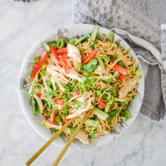 top view of sesame noodles with raw strips of cucumber, red bell peppers, and snap peas in a stone bowl with gold chopsticks resting on the rim all on a marble surface