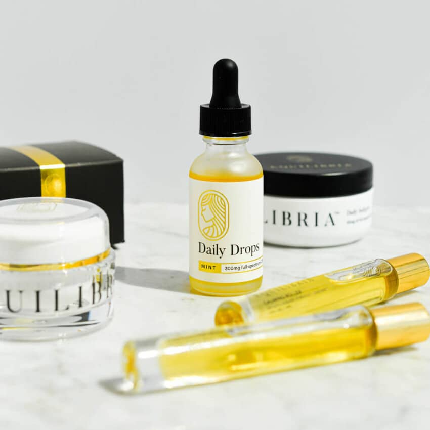 a glass bottle of equilibria cbd drops, rollers, relief cream, and softgels on a marble surface
